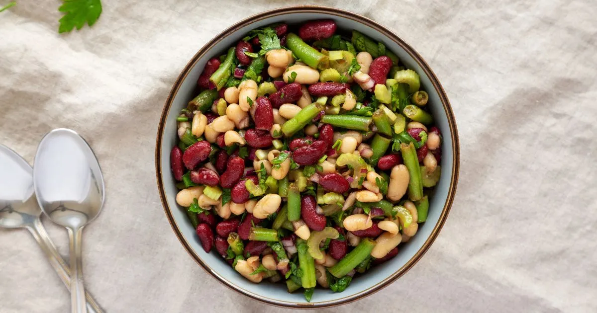 Dense bean salad recipe with colorful kidney beans, black beans, chickpeas, fresh tomatoes, cucumbers, red onions, and bell peppers, topped with a light vinaigrette and fresh herbs.